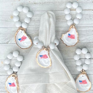 May include: Four white bead napkin rings with gold accents and oyster shell charms featuring an American flag design. The oyster shells are painted white with gold trim.