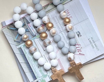 May include: A strand of wooden beads in a grey, white and gold colour scheme. The beads are strung on a white cord and have a distressed finish. The strand is topped with two gold crosses.