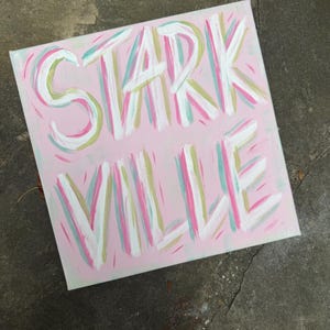 Op de afbeelding: Een vierkant canvas schilderij met een lichtroze achtergrond met het woord STARK VILLE handgeschilderd in wit, geaccentueerd met penseelstreken van lichtgroen, taling en donkerroze. De belettering is vet en gestileerd met zichtbare verftextuur.