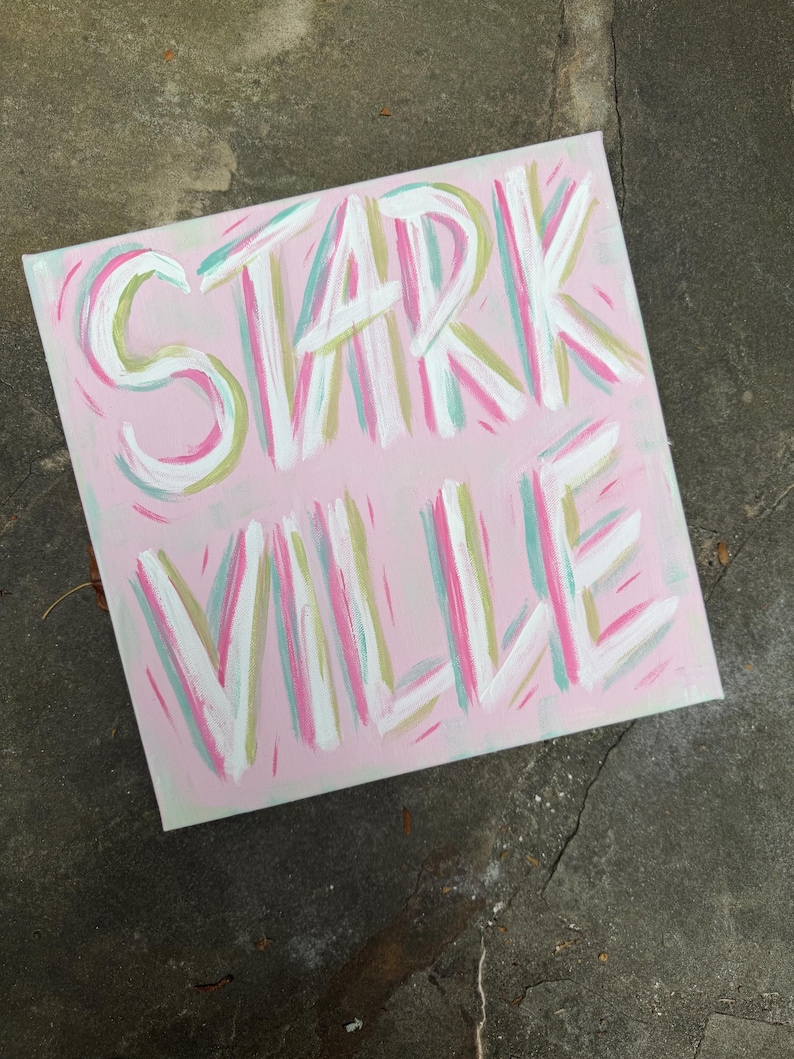 Op de afbeelding: Een vierkant canvas schilderij met een lichtroze achtergrond met het woord STARK VILLE handgeschilderd in wit, geaccentueerd met penseelstreken van lichtgroen, taling en donkerroze. De belettering is vet en gestileerd met zichtbare verftextuur.