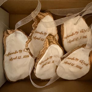 May include: A collection of oyster shell ornaments with gold edging and white interiors. Each shell is inscribed with the names "Amanda & Reid" and the date "April 25, 2026" in a delicate script, and attached with a sheer ribbon, presented in a box.