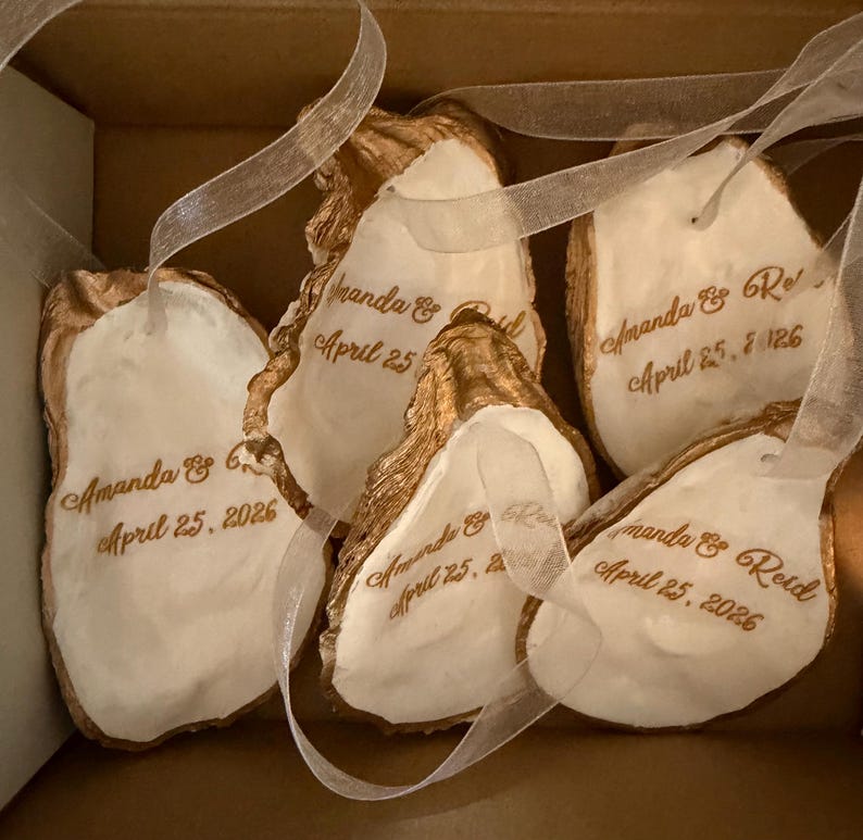 May include: A collection of oyster shell ornaments with gold edging and white interiors. Each shell is inscribed with the names "Amanda & Reid" and the date "April 25, 2026" in a delicate script, and attached with a sheer ribbon, presented in a box.