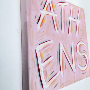 Op de afbeelding: Een vierkant schilderij op canvas met een lichtroze achtergrond waarop het woord ATHENS in grote witte letters is geschilderd. De letters zijn omlijnd met dunne, expressieve penseelstreken in tinten oranje, magenta, groenblauw en geel.
