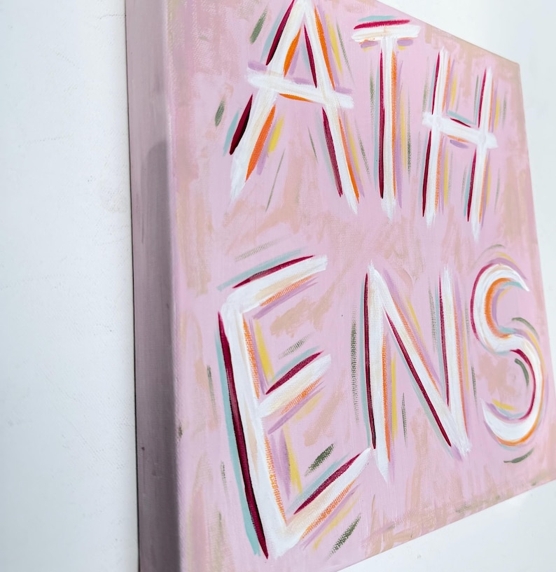 Op de afbeelding: Een vierkant schilderij op canvas met een lichtroze achtergrond waarop het woord ATHENS in grote witte letters is geschilderd. De letters zijn omlijnd met dunne, expressieve penseelstreken in tinten oranje, magenta, groenblauw en geel.