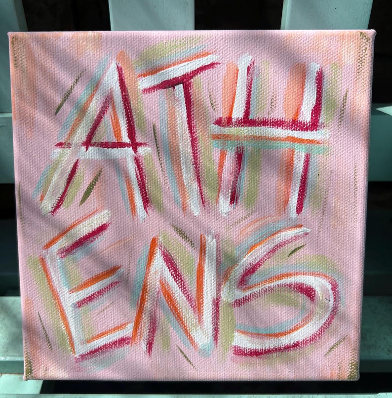 Peut inclure: Une peinture sur toile carr&eacute;e avec un fond rose clair pr&eacute;sentant le mot ATHENS peint en blanc, soulign&eacute; par des traits vibrants de magenta, d'orange et de vert clair. Le texte est superpos&eacute; sur un motif textur&eacute; de coups de pinceau multicolores.