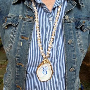 May include: A necklace featuring a strand of white and gold beads with a large oyster shell pendant. The shell has a gold-painted edge and a blue cursive letter E painted in the center. The necklace is worn over a blue and white striped shirt.