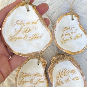 May include: Four oyster shells with gold edging and twine loops. Each shell features gold calligraphy with phrases such as "Today we said 'I Do!'" and "Thank you for being here!" Names and a date are also present.
