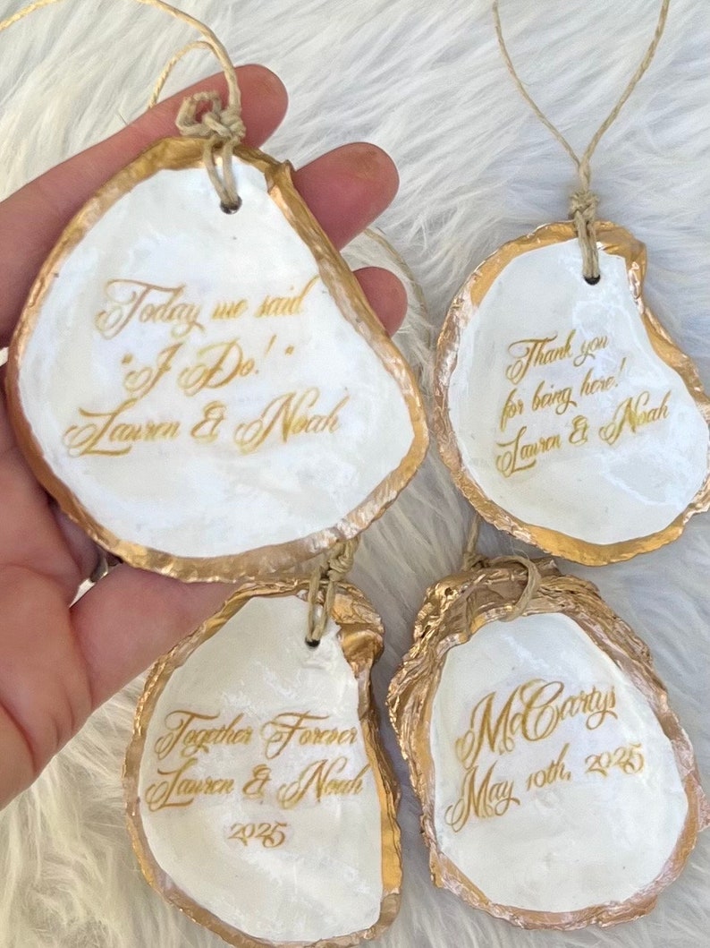May include: Four oyster shells with gold edging and twine loops. Each shell features gold calligraphy with phrases such as "Today we said 'I Do!'" and "Thank you for being here!" Names and a date are also present.