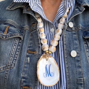 May include: A beaded necklace with large white and gold-accented beads. The necklace features a large, white oyster shell pendant with a gold rim and a blue letter "M". The necklace is worn over a blue and white striped shirt and a denim jacket.
