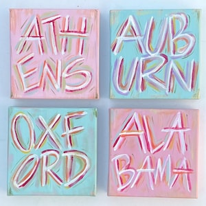 Peut inclure: Quatre toiles carr&eacute;es pr&eacute;sentant du texte peint &agrave; la main en blanc avec des contours au pinceau color&eacute;s. La toile en haut &agrave; gauche est rose avec le texte ATHENS, en haut &agrave; droite bleu clair avec AUBURN, en bas &agrave; gauche bleu clair avec OXFORD et en bas &agrave; droite rose avec ALABAMA.