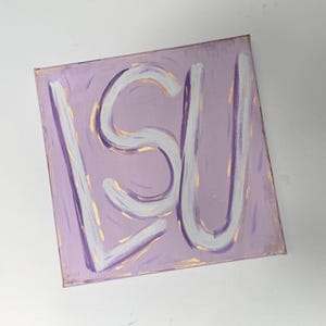 Op de afbeelding: Een vierkant canvas schilderij met de letters LSU in dikke witte verf tegen een lichtpaarse achtergrond. De letters zijn omlijnd met subtiele gouden penseelstreken en de paarse achtergrond bevat vage, verticale gouden verfaccenten.