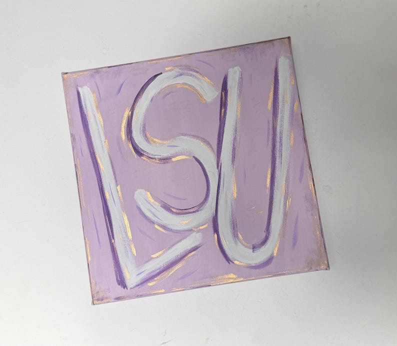 Op de afbeelding: Een vierkant canvas schilderij met de letters LSU in dikke witte verf tegen een lichtpaarse achtergrond. De letters zijn omlijnd met subtiele gouden penseelstreken en de paarse achtergrond bevat vage, verticale gouden verfaccenten.
