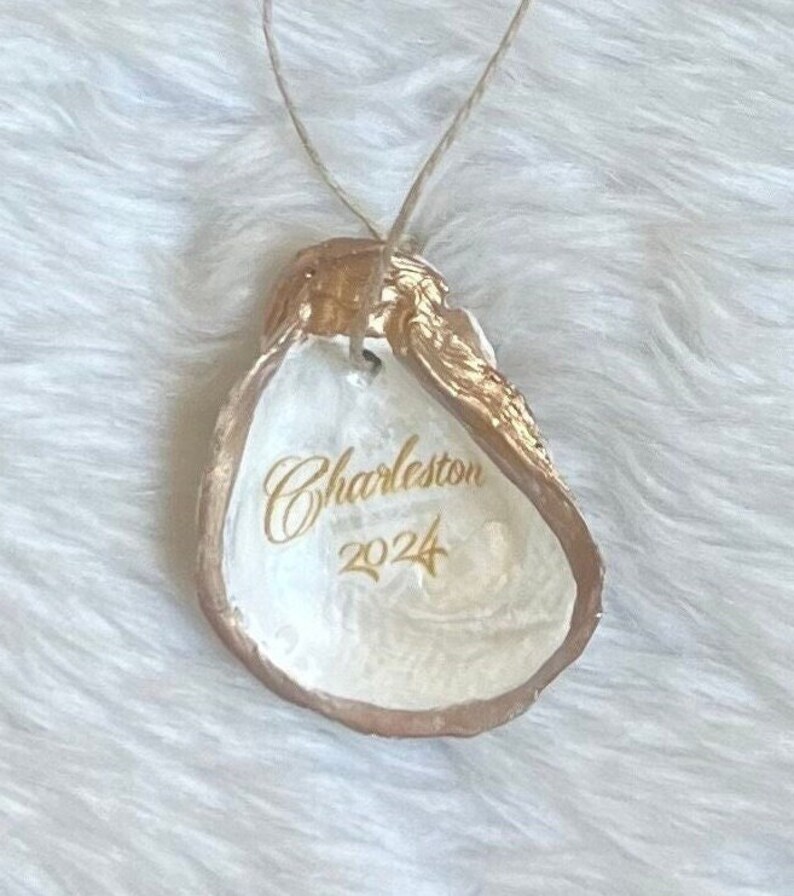 May include: An oyster shell ornament with gold edging and a hanging string. The interior is white with the word "Charleston" and the year "2024" in gold script. The shell is presented on a white, textured surface.