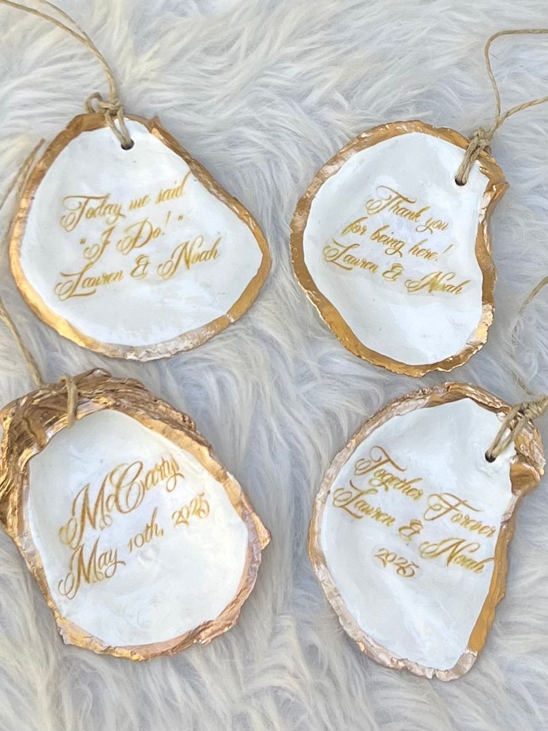 May include: Four white oyster shell ornaments with gold edging and twine. Each shell features gold calligraphy with phrases such as "Today we said I Do!" and "Thank you for being here!" for a wedding.