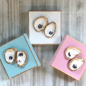 May include: Three square wooden boxes with gold trim, each painted a different colour: light blue, white, and pink. Each box features a pair of painted oyster shells with a pearl inside.