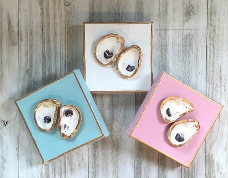 May include: Three square wooden boxes with gold trim, each painted a different colour: light blue, white, and pink. Each box features a pair of painted oyster shells with a pearl inside.