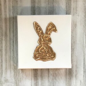 May include: A gold rabbit silhouette on a white canvas. The rabbit is made of a textured, raised material, giving it a 3D effect.