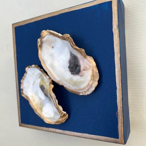 May include: Two oyster shells painted gold are mounted on a deep blue canvas with a gold border. The shells are arranged in a staggered position.