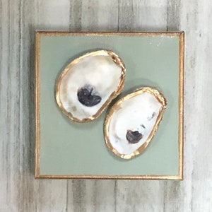 May include: Two oyster shells painted in white with gold accents on a sage green background. The shells are arranged in a horizontal line on a square canvas with a gold frame.