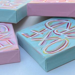 Peut inclure: Trois panneaux de toile rectangulaires peints en rose pastel et bleu clair. Les panneaux pr&eacute;sentent des lettres peintes &agrave; la main avec des coups de pinceau blancs, roses et jaunes. Le texte sur les toiles inclut les mots "LOVE", "KIND" et "DREAMS".