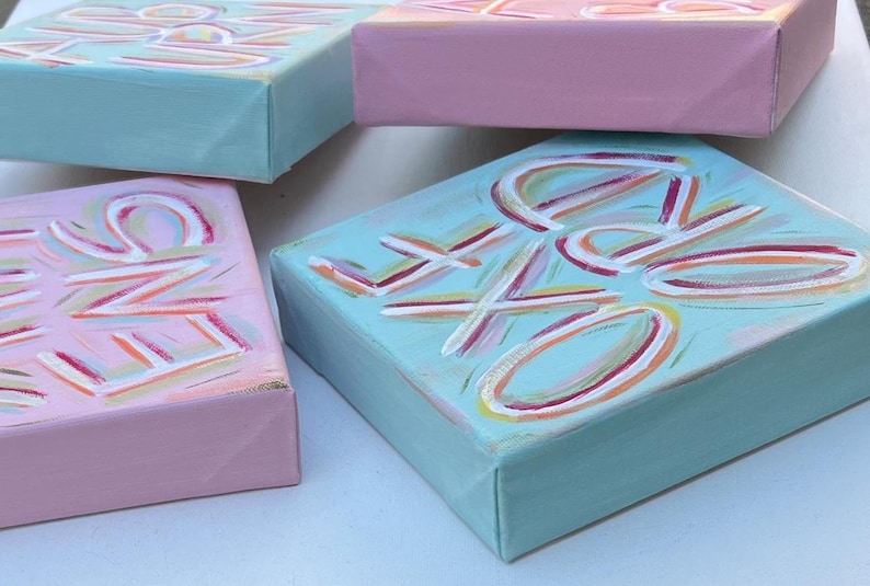 Peut inclure: Trois panneaux de toile rectangulaires peints en rose pastel et bleu clair. Les panneaux pr&eacute;sentent des lettres peintes &agrave; la main avec des coups de pinceau blancs, roses et jaunes. Le texte sur les toiles inclut les mots "LOVE", "KIND" et "DREAMS".