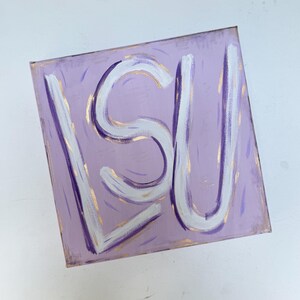Peut inclure: Une peinture sur toile carr&eacute;e pr&eacute;sentant un fond violet clair avec les lettres LSU peintes en coups de pinceau blancs &eacute;pais. L'&oelig;uvre comprend de subtils accents de peinture dor&eacute;e et de petits traits verticaux violets et dor&eacute;s dispers&eacute;s.