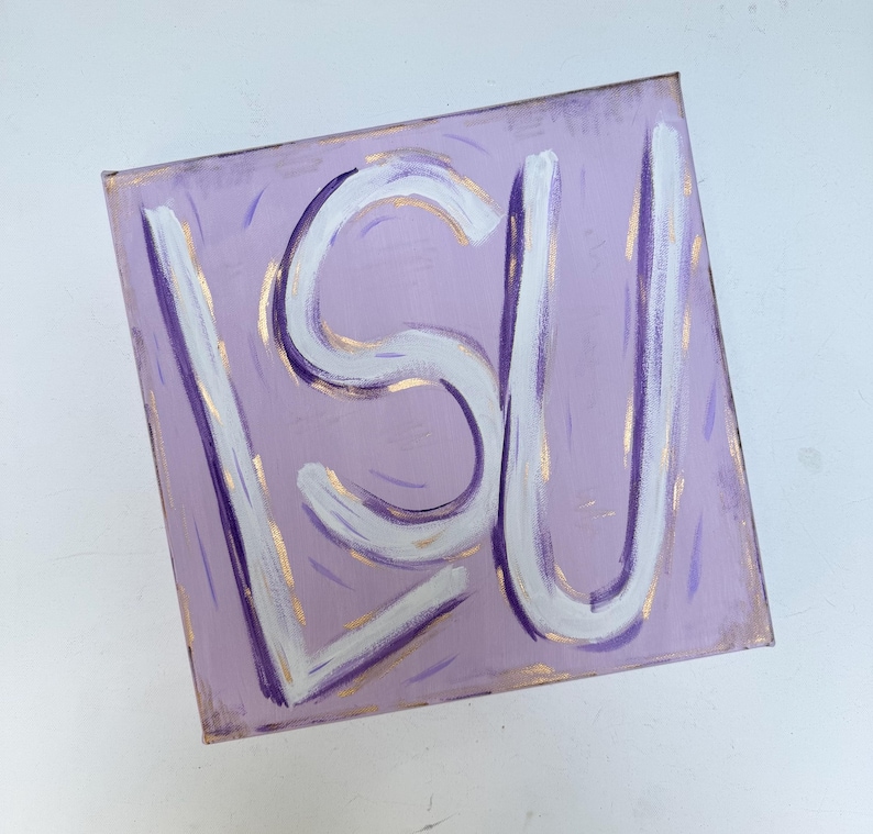 Peut inclure: Une peinture sur toile carr&eacute;e pr&eacute;sentant un fond violet clair avec les lettres LSU peintes en coups de pinceau blancs &eacute;pais. L'&oelig;uvre comprend de subtils accents de peinture dor&eacute;e et de petits traits verticaux violets et dor&eacute;s dispers&eacute;s.