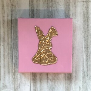 May include: A pink canvas with a gold, abstract, stylized figure of a person in a dress. The figure is raised from the canvas.
