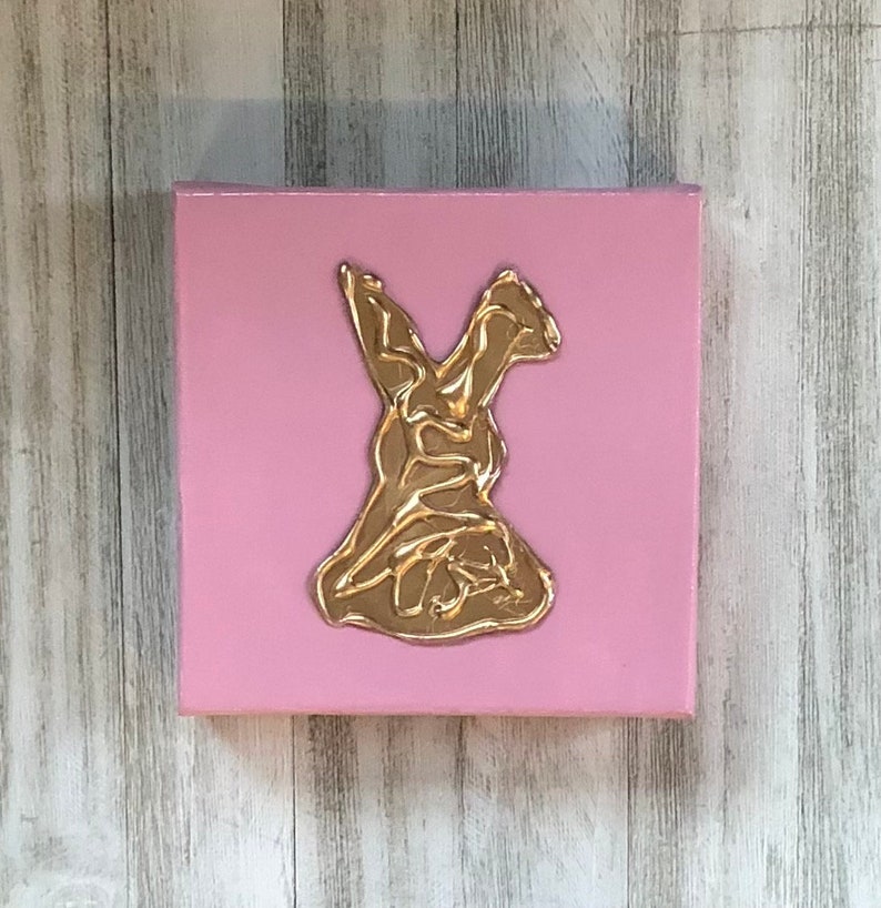 May include: A pink canvas with a gold, abstract, stylized figure of a person in a dress. The figure is raised from the canvas.
