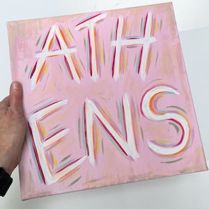 Op de afbeelding: Een vierkant canvas schilderij met een lichtroze achtergrond waarop het woord ATHENS in grote witte letters is geschilderd. De letters zijn omlijnd met dunne, kleurrijke penseelstreken in tinten oranje, magenta en taling, wat een levendig effect cre&euml;ert.