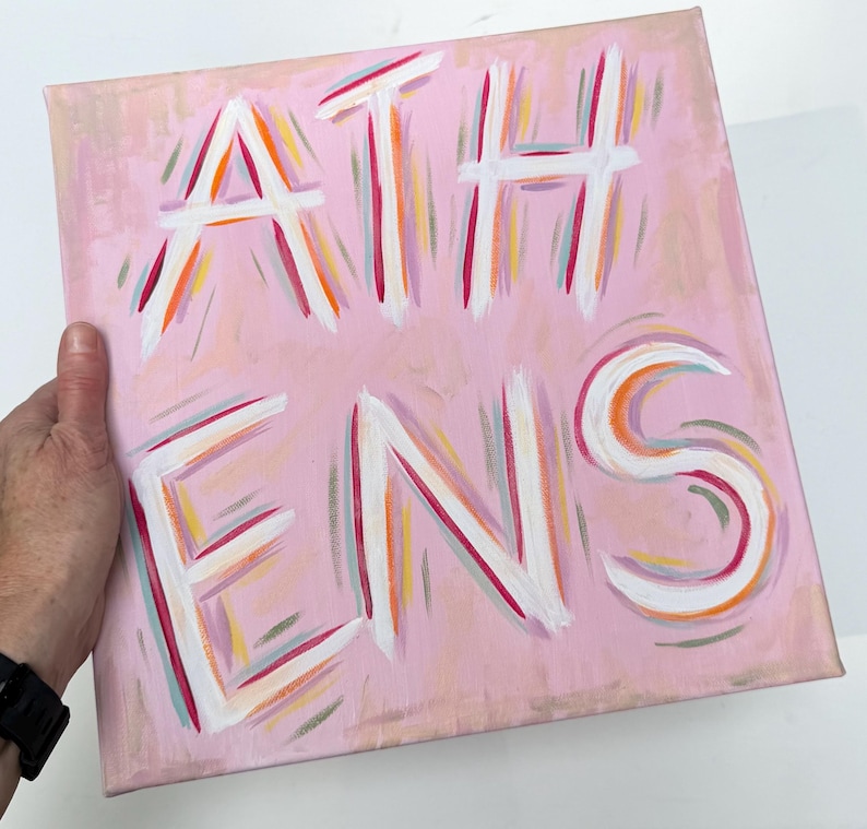 Op de afbeelding: Een vierkant canvas schilderij met een lichtroze achtergrond waarop het woord ATHENS in grote witte letters is geschilderd. De letters zijn omlijnd met dunne, kleurrijke penseelstreken in tinten oranje, magenta en taling, wat een levendig effect cre&euml;ert.