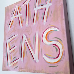 Op de afbeelding: Een vierkant canvas schilderij met een lichtroze achtergrond en het woord ATHENS met de hand geschilderd in grote, dikke witte letters. De letters zijn geaccentueerd met dunne, expressieve penseelstreken in tinten oranje, rood en blauwgroen.