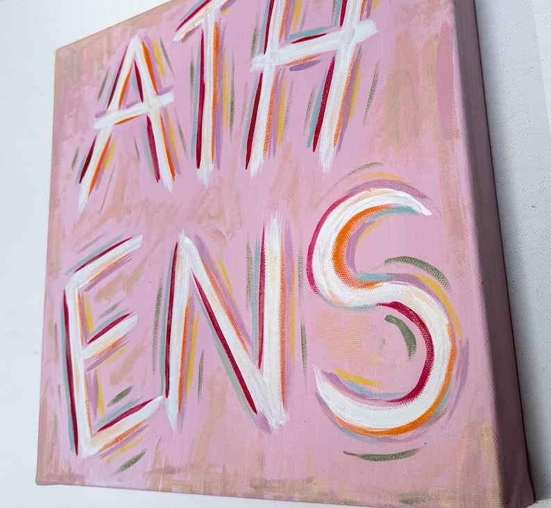 Op de afbeelding: Een vierkant canvas schilderij met een lichtroze achtergrond en het woord ATHENS met de hand geschilderd in grote, dikke witte letters. De letters zijn geaccentueerd met dunne, expressieve penseelstreken in tinten oranje, rood en blauwgroen.