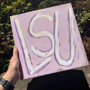 Op de afbeelding: Een vierkant canvas schilderij met de letters LSU geschilderd in dikke witte penseelstreken met gouden accenten. De achtergrond is een licht lavendelkleur met subtiele paarse en gouden verfstreken die een textuur op het oppervlak cre&euml;ren.