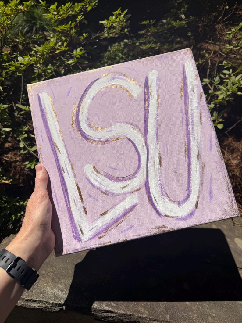 Op de afbeelding: Een vierkant canvas schilderij met de letters LSU geschilderd in dikke witte penseelstreken met gouden accenten. De achtergrond is een licht lavendelkleur met subtiele paarse en gouden verfstreken die een textuur op het oppervlak cre&euml;ren.
