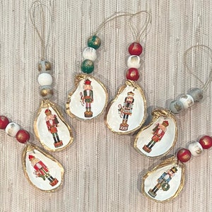 May include: Six Christmas ornaments featuring nutcracker figures painted on oyster shells. Each ornament is strung with beads in red, white, and green.