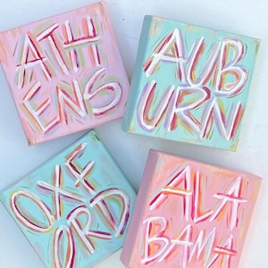 Peut inclure: Quatre toiles carr&eacute;es pr&eacute;sentant des lettres peintes &agrave; la main en blanc avec des contours color&eacute;s. La toile en haut &agrave; gauche est rose avec le texte ATHENS, celle en haut &agrave; droite est bleu clair avec AUBURN, en bas &agrave; gauche bleu clair avec OXFORD, et en bas &agrave; droite rose avec ALABAMA.