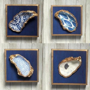 May include: Four square blue and gold framed canvases with white oyster shells painted with blue and white patterns. The patterns include a geometric design, a fish design, a zig-zag design, and a solid white design.