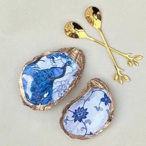 May include: Two gold oyster shell dishes with blue and white floral designs. One dish features a peacock, the other has a floral pattern. The dishes are paired with two gold spoons with leaf-shaped handles.