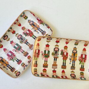 May include: Two rectangular serving trays with gold trim feature a white background with a repeating pattern of nutcracker figurines in various colors and poses.