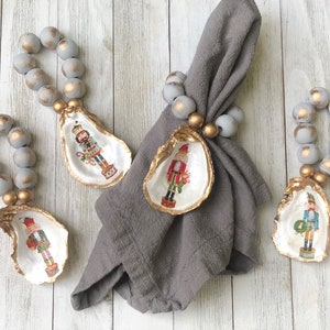 May include: Four gray and gold beaded napkin rings with a nutcracker design on a white oyster shell. The napkin rings are hanging from the shells.