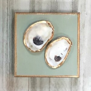 May include: Two oyster shells painted with gold leaf on a sage green background. The shells are arranged in a vertical orientation.
