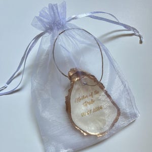 May include: An oyster shell pendant with gold edging, inscribed with "Mother of the Bride" and the date "09.07.2024". The pendant is suspended from a silver wire and presented in a sheer, pale purple drawstring bag.