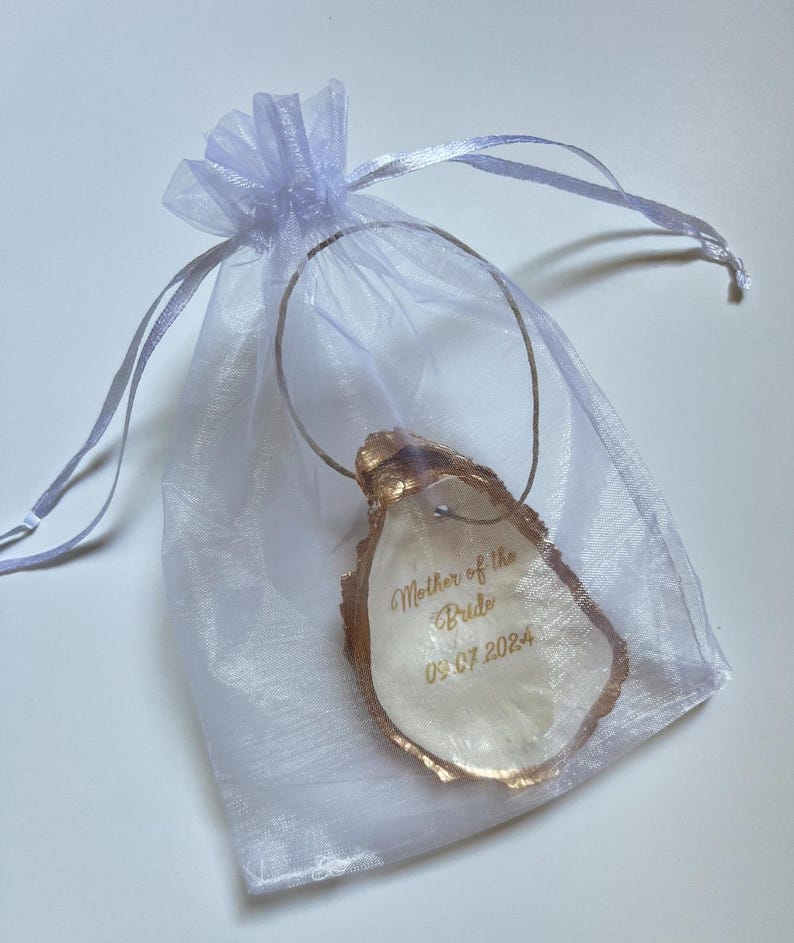 May include: An oyster shell pendant with gold edging, inscribed with "Mother of the Bride" and the date "09.07.2024". The pendant is suspended from a silver wire and presented in a sheer, pale purple drawstring bag.