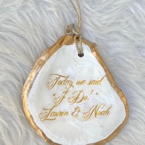 May include: An oyster shell ornament with a gold border and white interior. The shell is inscribed with the words "Today we said 'I Do!' Lauren & Noah" in gold script. A twine loop is attached for hanging.