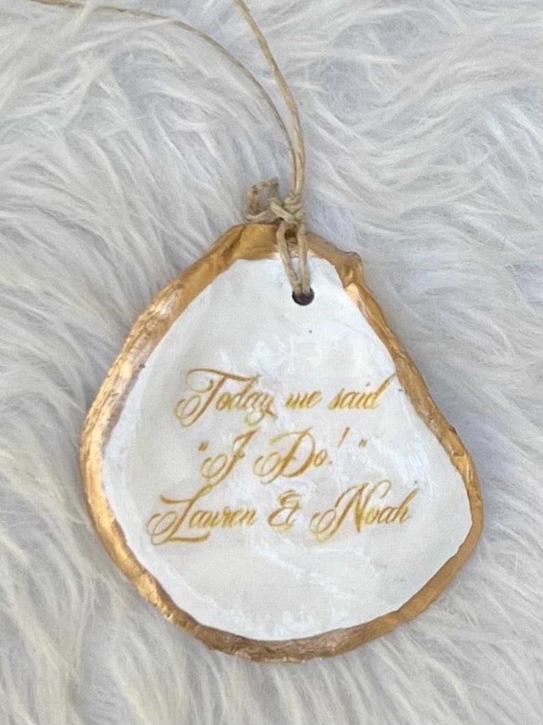 May include: An oyster shell ornament with a gold border and white interior. The shell is inscribed with the words "Today we said 'I Do!' Lauren & Noah" in gold script. A twine loop is attached for hanging.