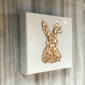 May include: A white canvas with a gold rabbit design. The rabbit is outlined in gold and is in a sitting position.