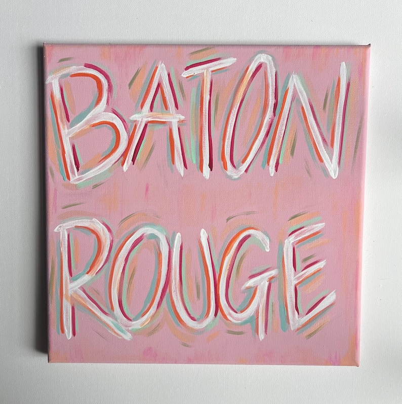 Peut inclure: Une peinture sur toile carr&eacute;e avec un fond rose clair pr&eacute;sentant les mots BATON ROUGE peints &agrave; la main en blanc. Les lettres sont soulign&eacute;es par des coups de pinceau superpos&eacute;s dans des tons orange, bleu sarcelle et rose fonc&eacute;.