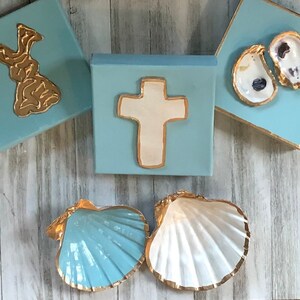 May include: Three blue square canvases with gold trim, each featuring a different design. The first canvas has a gold bunny rabbit, the second has a white cross, and the third has two gold-trimmed oyster shells.