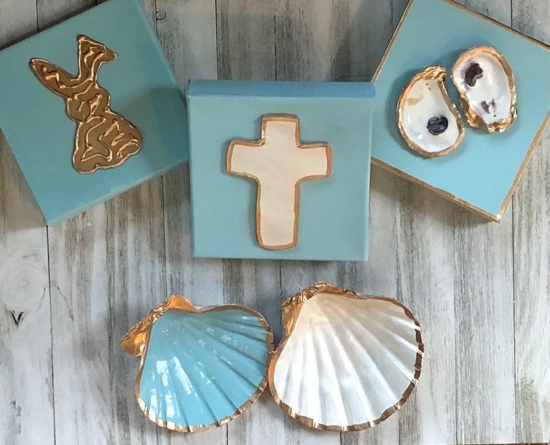May include: Three blue square canvases with gold trim, each featuring a different design. The first canvas has a gold bunny rabbit, the second has a white cross, and the third has two gold-trimmed oyster shells.